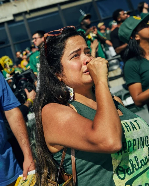 A woman in a green Oakland shirt watches a sporting event with fans; she has long hair and sunglasses on her head.