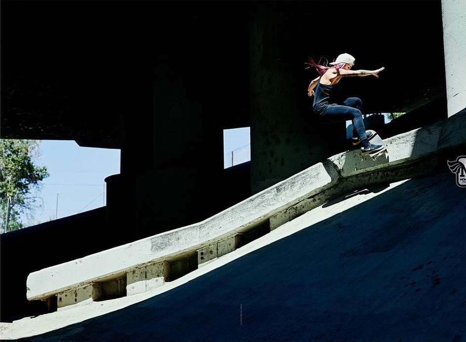 A woman performs a skateboard trick on a concrete embankment ramp under a bridge, wearing a black top, jeans, and a white baseball cap.
