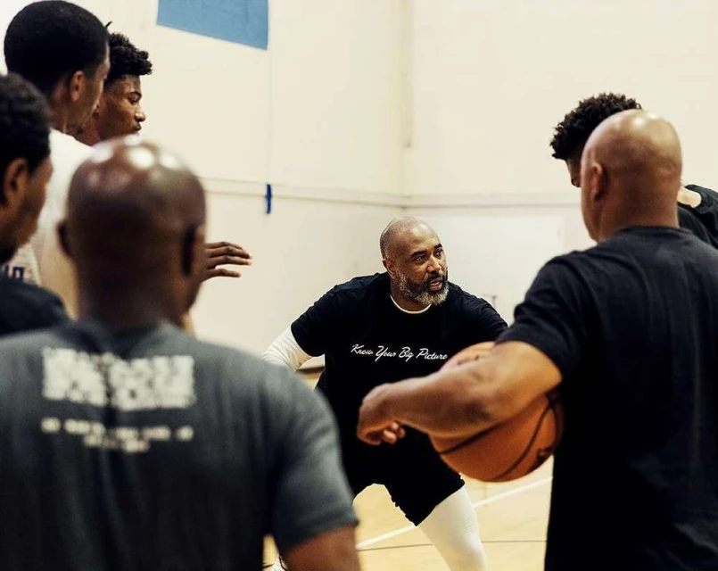 Indoor shot of a basketball coach with a beard, standing among a group of young African American male athletes on a gymnasium court.