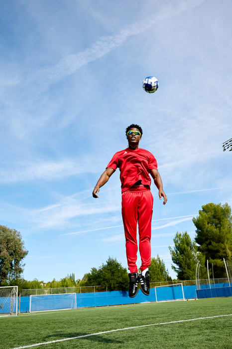 A man in red sportswear leaps to head a soccer ball on a sunny green field with a blue sky and trees in the background.