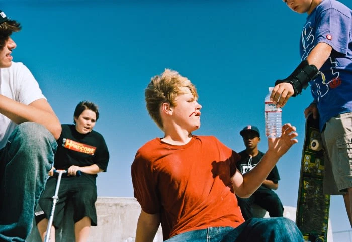 Group of teenage boys outside on sunny day, one holding water bottle, another with skateboard and wrist brace, blue sky background.