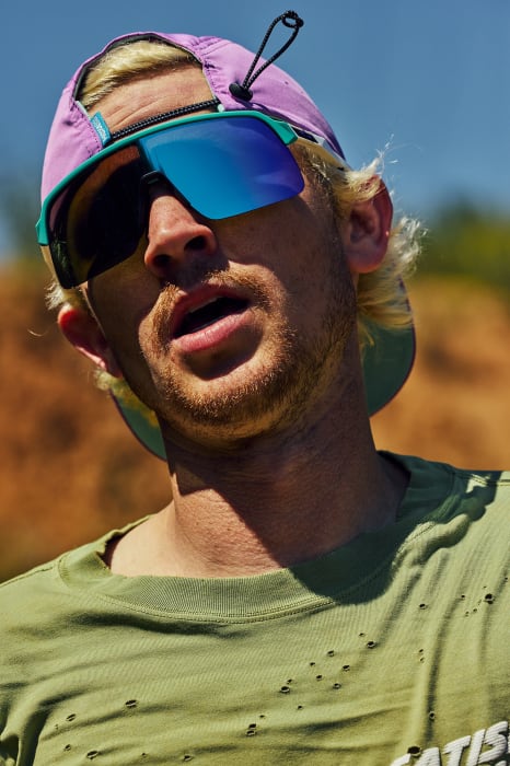 Outdoor portrait of a blond adult male wearing sunglasses and baseball cap, posed in medium shot under sunlight, with blurred background.