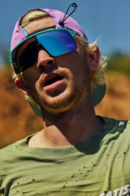 Outdoor portrait of a blond adult male wearing sunglasses and baseball cap, posed in medium shot under sunlight, with blurred background.