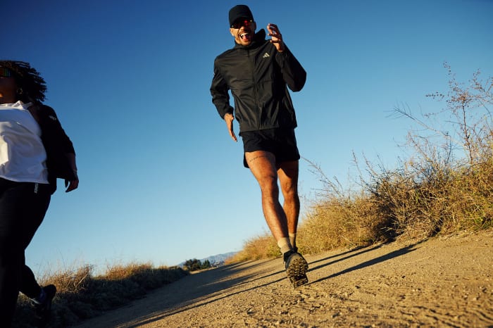 A man in athletic wear runs on a dirt path with another person in the background under a clear blue sky during the daytime.