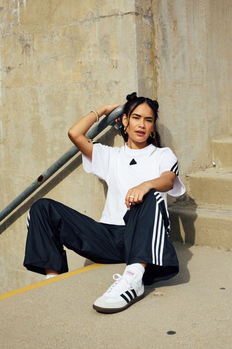 A woman wearing a white Adidas T-shirt and black track pants sits on concrete steps in bright sunlight. She is posing and smiling.