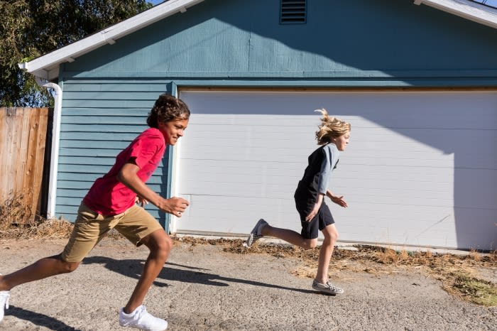 Two children race each other on an asphalt surface in front of a light blue building with a white garage door on a sunny day.