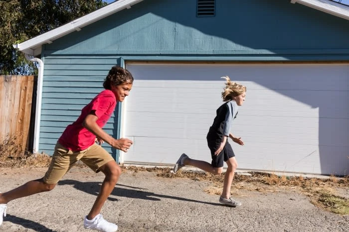 Two children race each other on an asphalt surface in front of a light blue building with a white garage door on a sunny day.