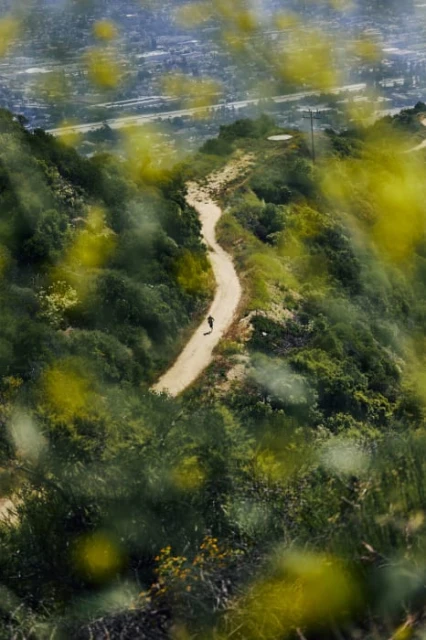 Person hikes a winding dirt trail through green hills on a sunny day, with a blurred cityscape visible in the background.