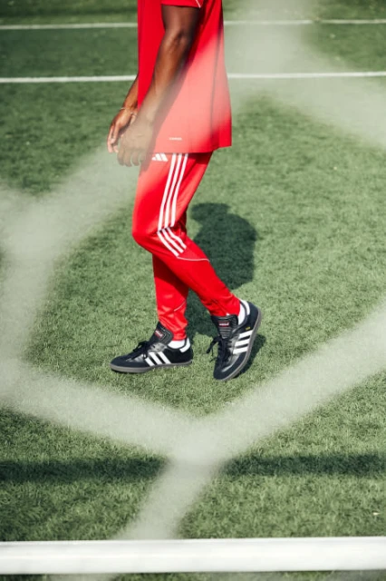 An adult male in red athletic wear and Adidas Samba shoes stands on a green sports field behind a white net, showcasing a sporty lifestyle.