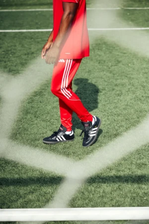 An adult male in red athletic wear and Adidas Samba shoes stands on a green sports field behind a white net, showcasing a sporty lifestyle.