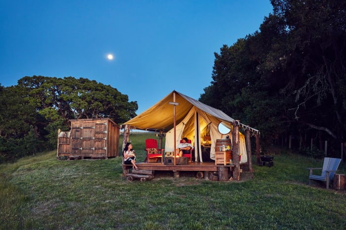 Glamping tent at dusk on grassy hill with two people outside, lit by the moon, with chairs, small table, and wooden shed.