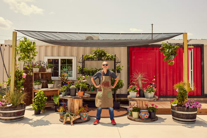 Man stands in front of a vibrant flower shop. He is wearing an apron and sunglasses, with many plants and flowers surrounding him.