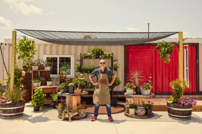 Man stands in front of a vibrant flower shop. He is wearing an apron and sunglasses, with many plants and flowers surrounding him.