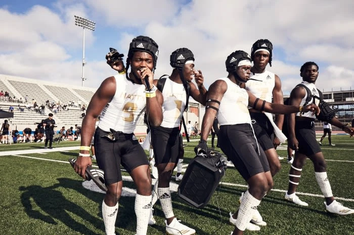 A group of six athletic adult men wearing football gear outdoors on a sunny day on a football field with stadium seating in the background.
