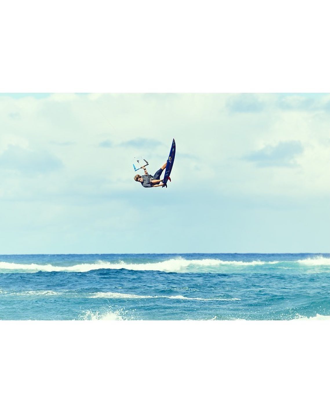 A kitesurfer performs an aerial stunt, twisting in the air above the ocean with a blue kiteboard, on a sunny day with a blue sky backdrop.