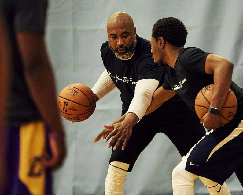 Two adult males in black t-shirts dribble basketballs in an indoor gym setting, focusing on training and athletic skill development.