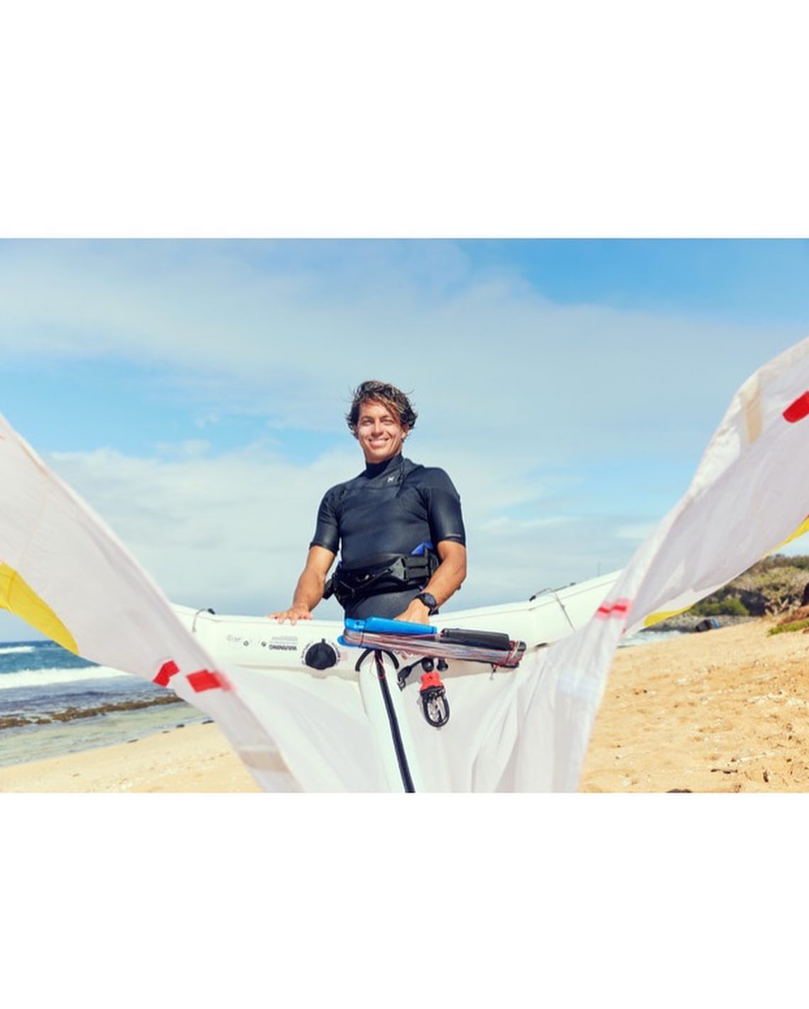 Smiling man in wetsuit holding kite bar on sandy beach with blue sky and ocean backdrop, enjoying watersport activity.