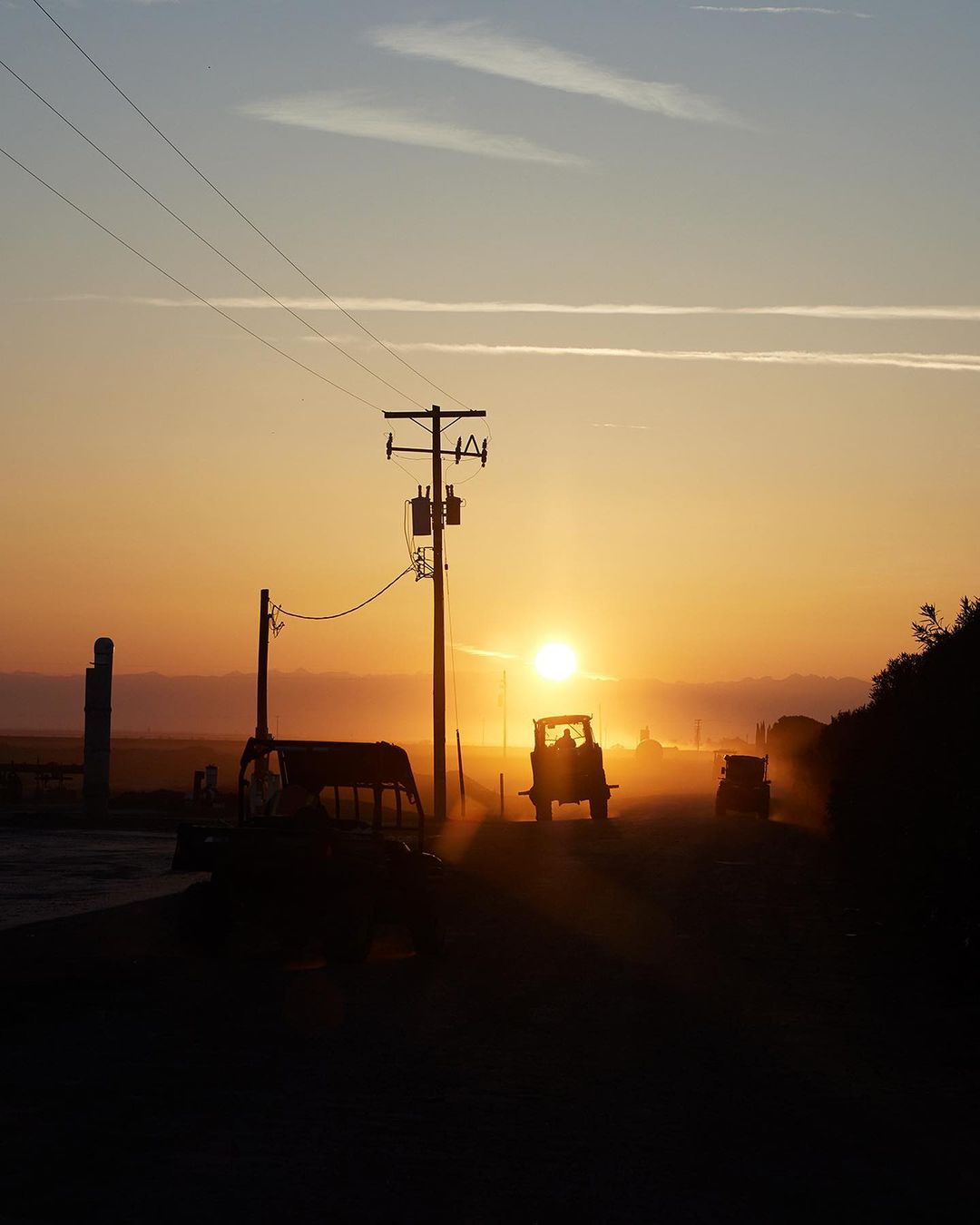 Silhouette of tractors driving on a rural road at sunset with power lines overhead. Silhouetted machinery and setting sun.