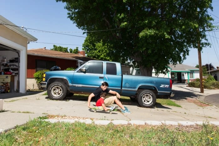 A blue pickup truck parked in front of a house with two people, one adult male and one child, sitting on a skateboard in the driveway.