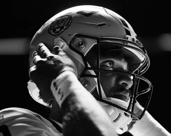 Black and white shot of a football player in uniform wearing a helmet. The focus is on the athlete's focused expression.