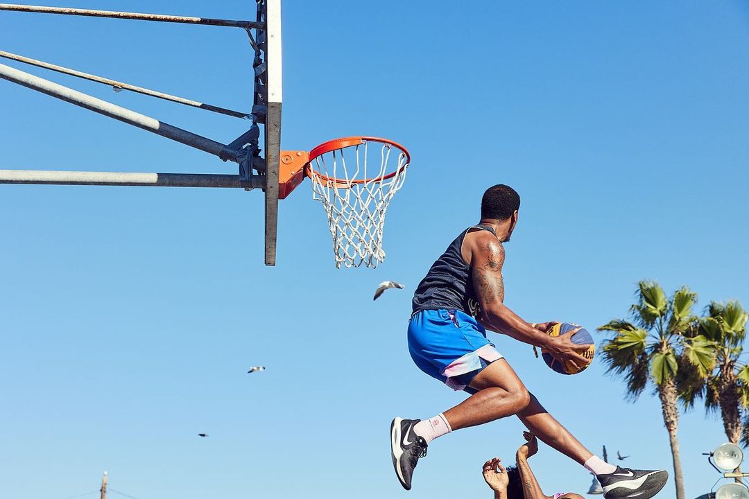 A male basketball player in mid-air with a ball and sneakers, with a basketball hoop, palm trees, and blue sky background on a sunny day.