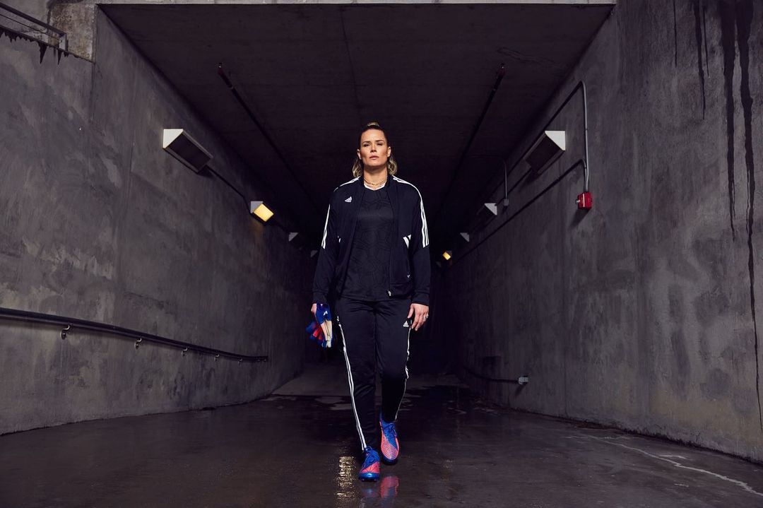Adult woman in black track suit walking through a concrete tunnel, holding running shoes, illuminated by overhead lights.