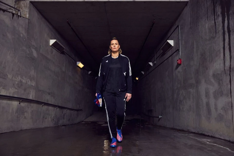 Adult woman in black track suit walking through a concrete tunnel, holding running shoes, illuminated by overhead lights.