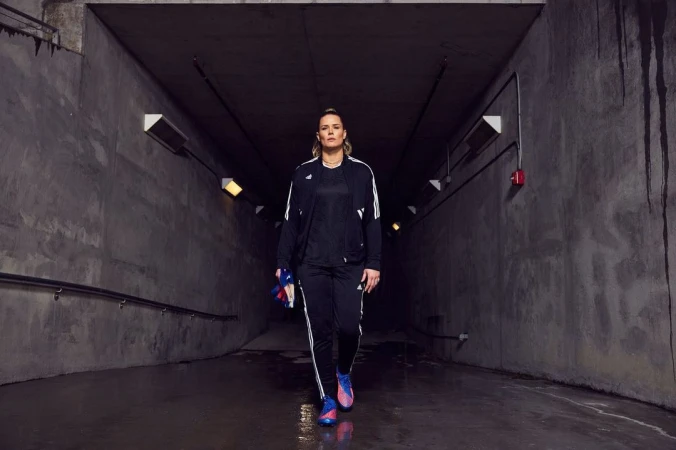 Adult woman in black track suit walking through a concrete tunnel, holding running shoes, illuminated by overhead lights.