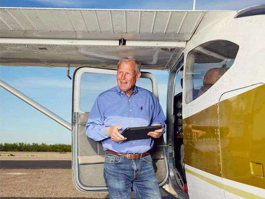 An older adult man stands near a small airplane on an airfield, holding a tablet, with a second person visible inside the plane.