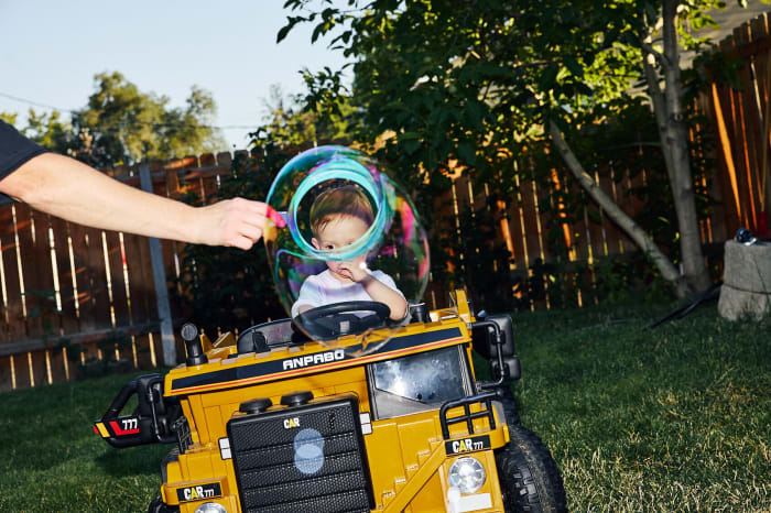 Toddler in a toy bulldozer with bubbles in an outdoor grassy area with trees and a wooden fence, creating a playful scene.