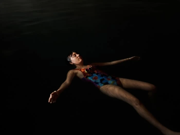 Woman in a floral swimsuit floats in dark water with arms outstretched, eyes closed, captured in a medium shot emphasizing tranquility.
