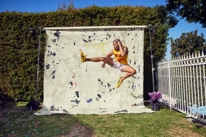 Athletic man in yellow tank top and white shorts mid-air with a kick against a painted backdrop in a grassy outdoor setting.