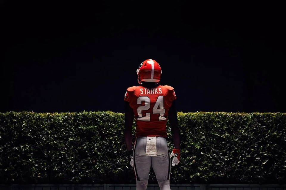American football player in full uniform, number 24, stands facing away from the viewer in front of a green hedge.