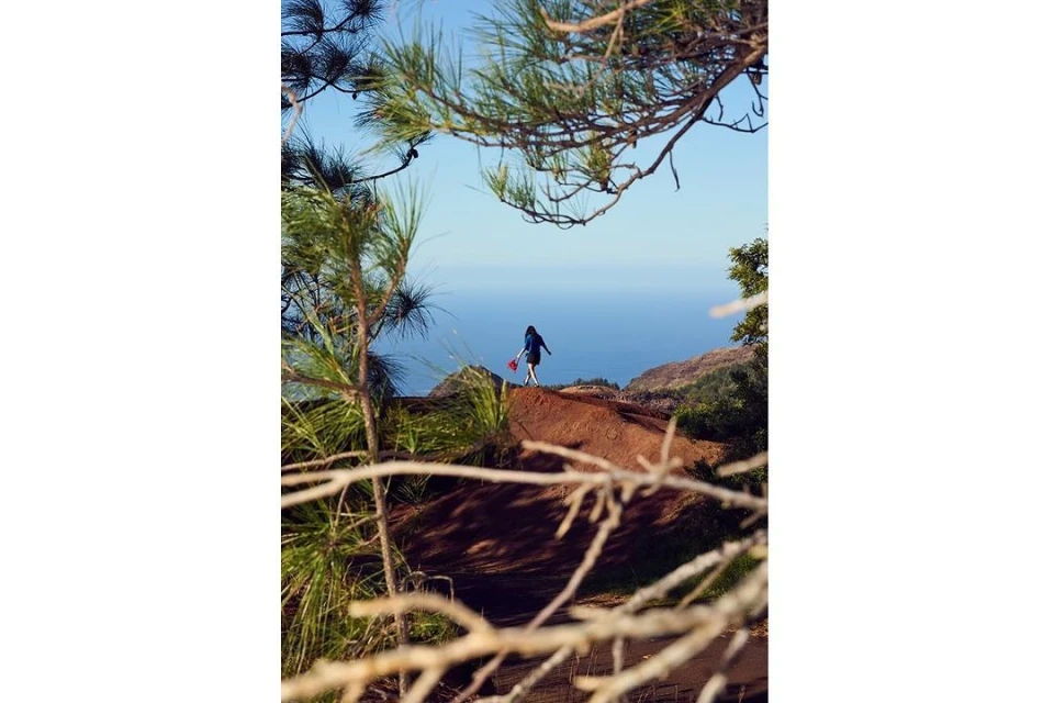Woman standing on a red dirt hill with a red object in hand, framed by green trees and a distant blue ocean under a clear sky.