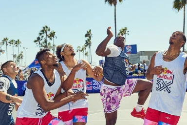 Five male basketball players in sportswear looking up intensely on an outdoor court with palm trees.