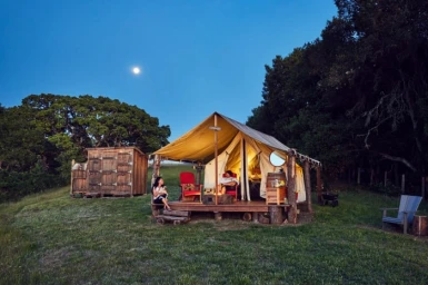 Large canvas tent set up on a wooden deck in a grassy field with a wooden outhouse and a moonlit sky at dusk.