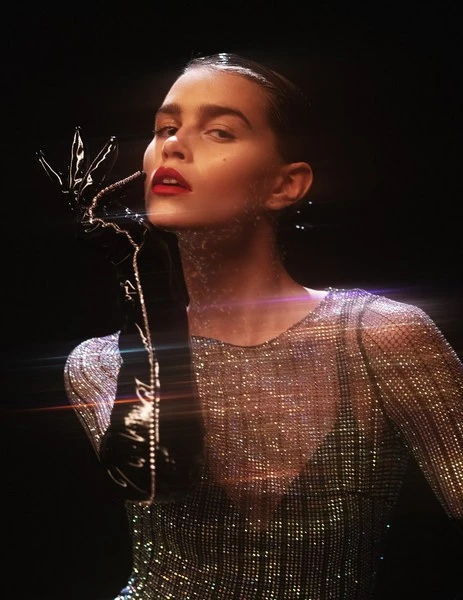 Elegant portrait of an adult woman with dark hair, red lipstick, and long black gloves, wearing a sparkling top, against a dark background.