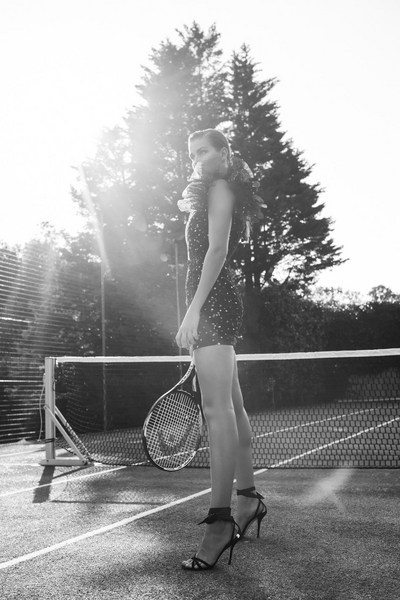 Monochrome shot of a woman in a romper and heels holding a tennis racket on a court, trees and sunlight in the background.