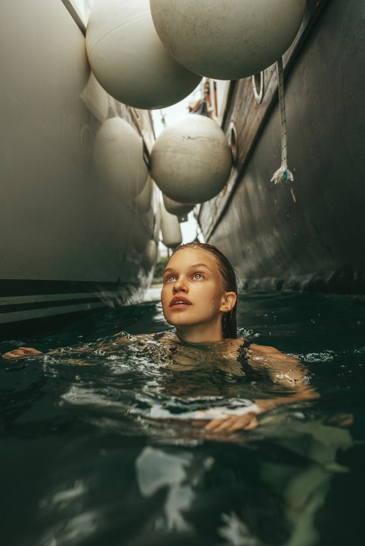 Woman in water between boats looks up at the white buoys hanging above on a cloudy day with cool toned lighting and focus on the model.