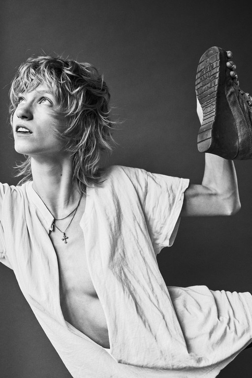 Black and white portrait of a person with blonde hair, wearing a white shirt, posing with a boot held overhead in a studio setting.