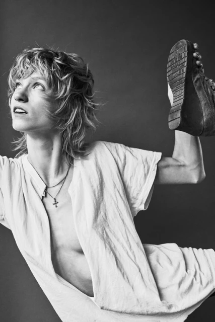 Black and white portrait of a person with blonde hair, wearing a white shirt, posing with a boot held overhead in a studio setting.