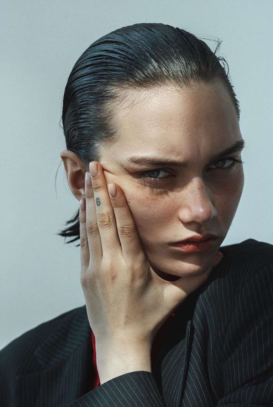 Close-up portrait of a young woman with hand on face, wet hair, and pinstripe blazer against a neutral background in studio lighting.