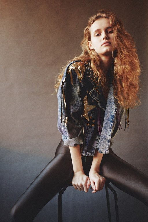 A young woman with curly red hair poses sitting on a stool, wearing black leggings and a denim jacket with gold decorations.