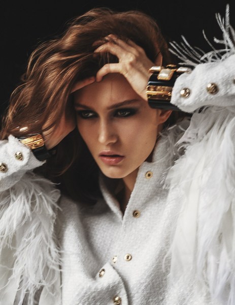 Close-up fashion portrait of a woman in a white jacket with feather trim and large gold cuff bracelets against a dark background.