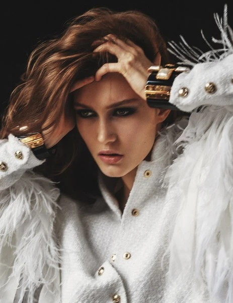 Close-up fashion portrait of a woman in a white jacket with feather trim and large gold cuff bracelets against a dark background.