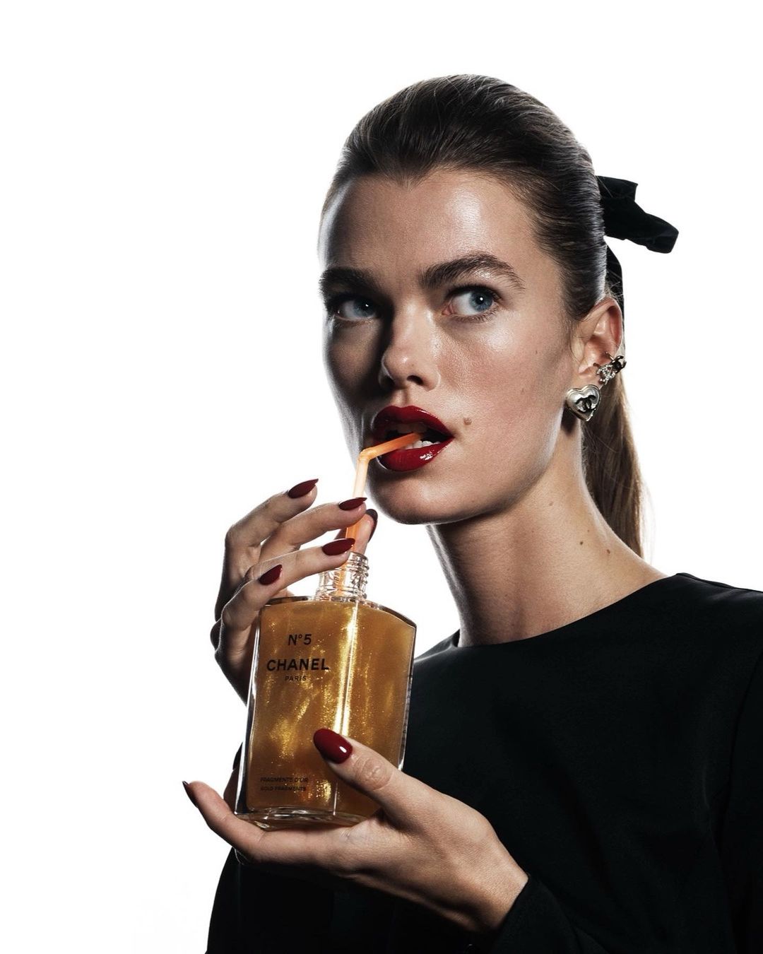 Woman with red lips holds Chanel No. 5 gold body oil bottle with a straw. Studio shot against white background. Beauty advertising.