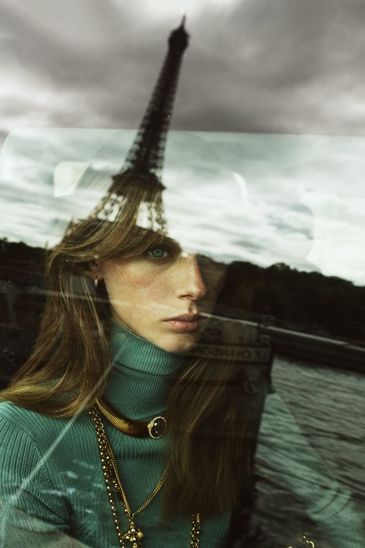 Woman wearing a turtleneck and gold jewelry with the Eiffel Tower reflected in glass, creating a surreal and fashionable portrait.