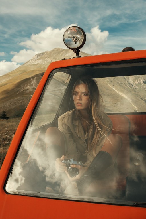 Woman with camera sits in a red truck with a mirror on top, mountain and sky background, creating a lifestyle editorial scene.