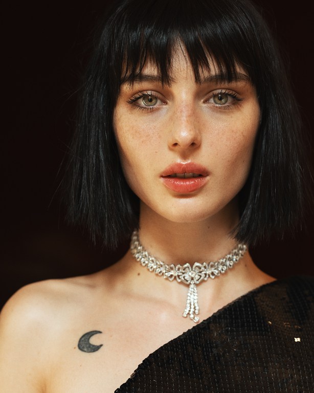 Close-up portrait of a woman with dark hair and freckles wearing a jeweled necklace and a black sequin top against a dark background.