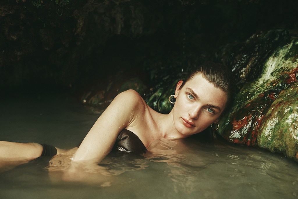 Adult woman with gold hoop earrings in black swimwear lies in clear water near a rocky outcrop inside a dark, natural rock cave.